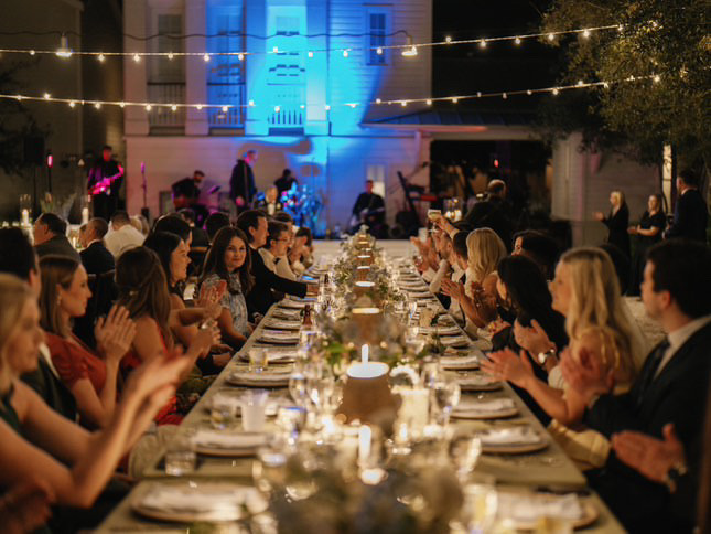 Nighttime outdoor banquet with elegant tableware, candles, string lights, and guests enjoying live music.