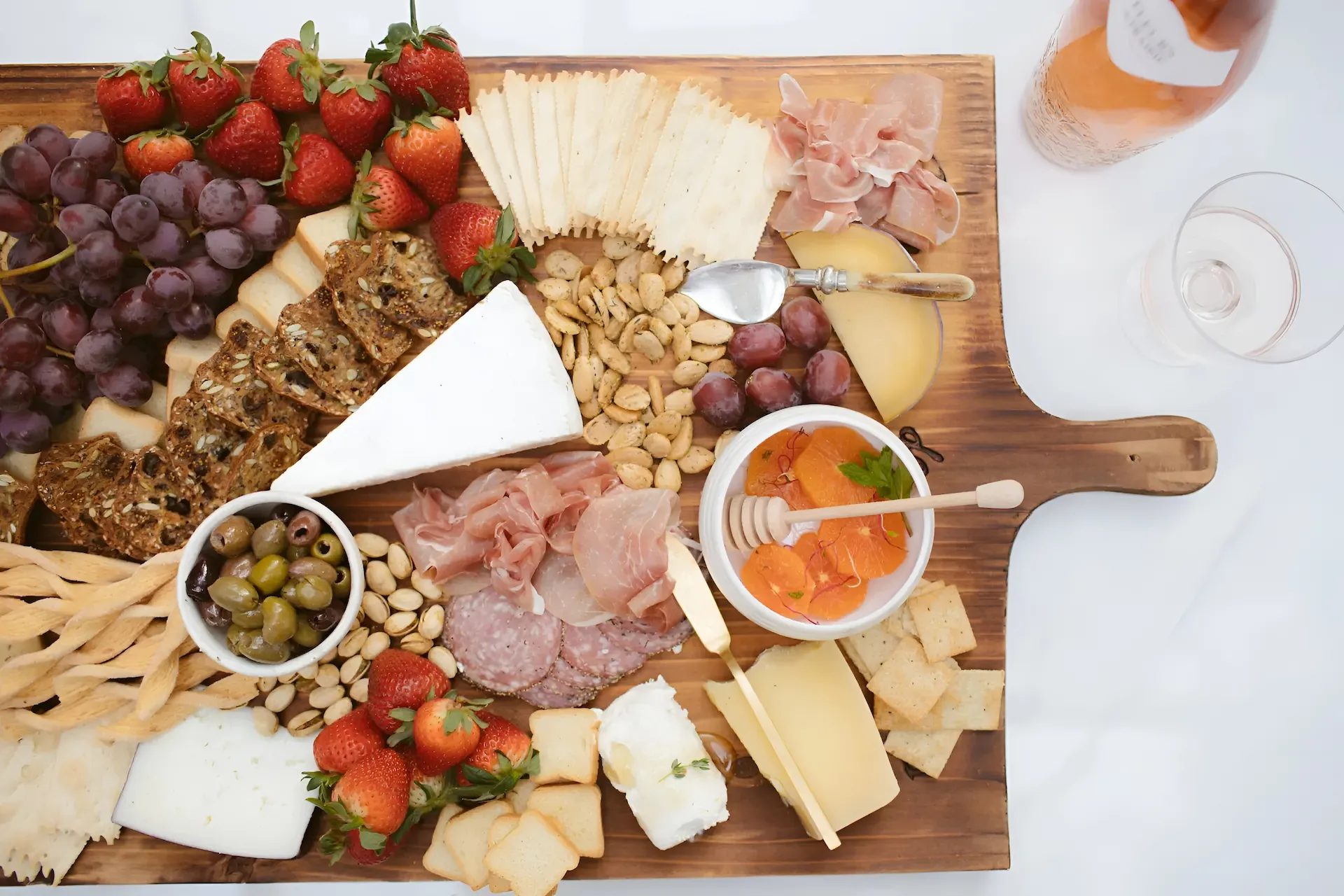 Charcuterie board with fruits, cheeses, cured meats, nuts, olives, crackers, and a glass of rosé wine.